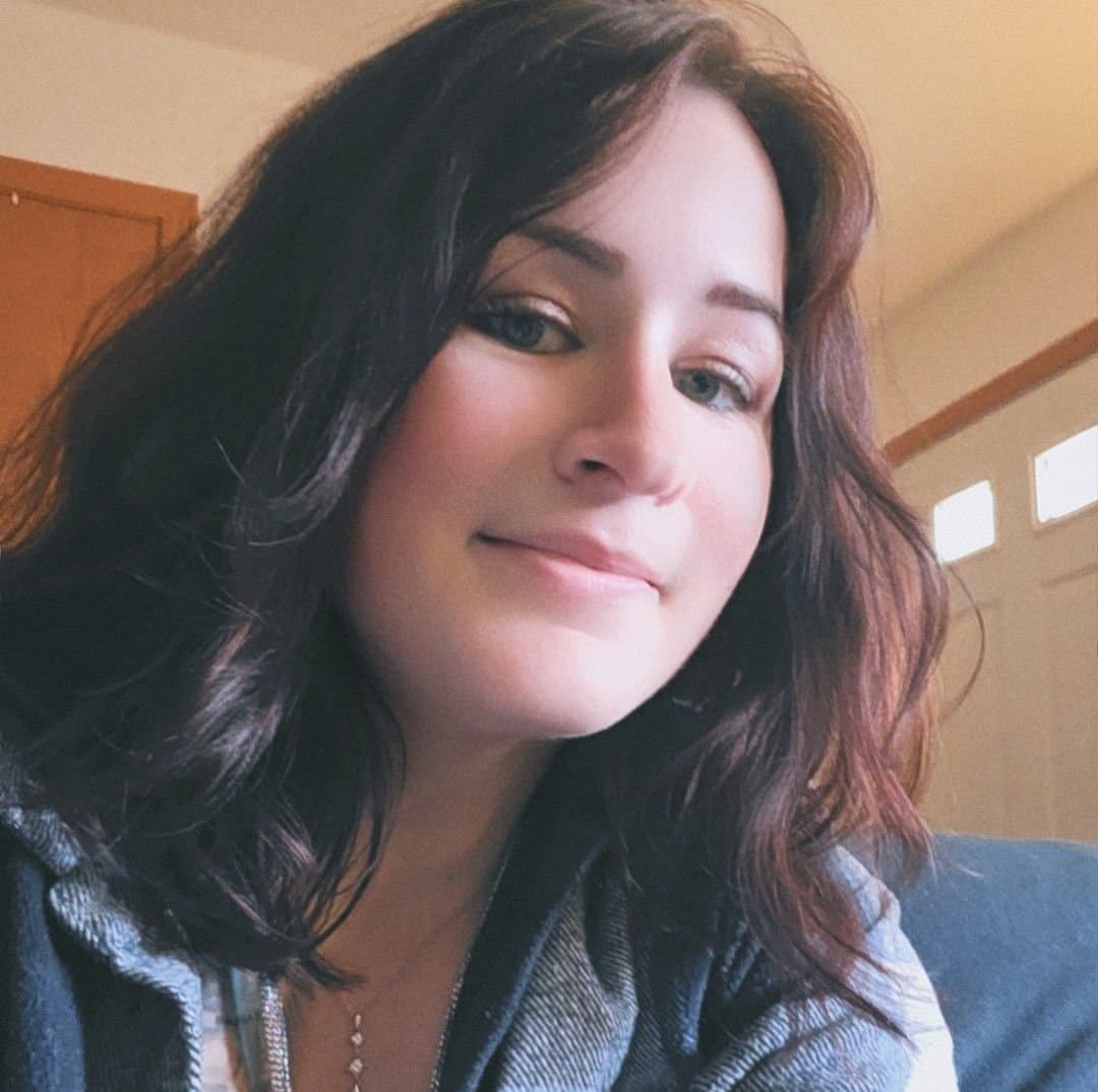 A young woman with dark wavy hair smiles gently, wearing a grey jacket indoors.